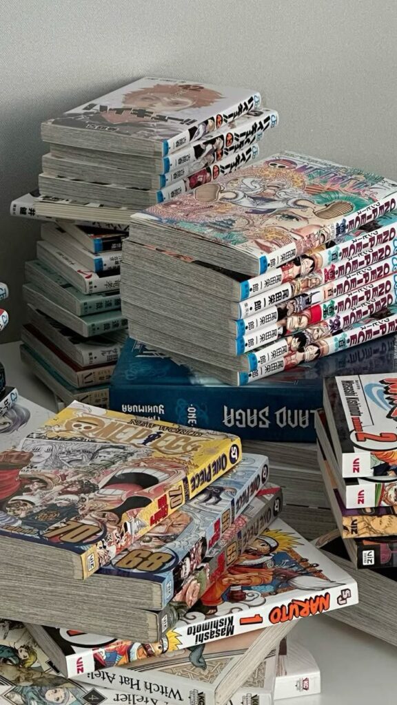 Stacks of various manga volumes on a desk, featuring titles like One Piece and Naruto.