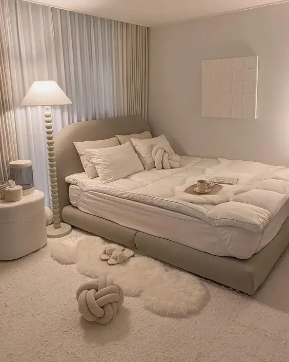 Cozy neutral bedroom with plush bedding, modern décor, and a warm floor lamp creating a serene, minimalist atmosphere.