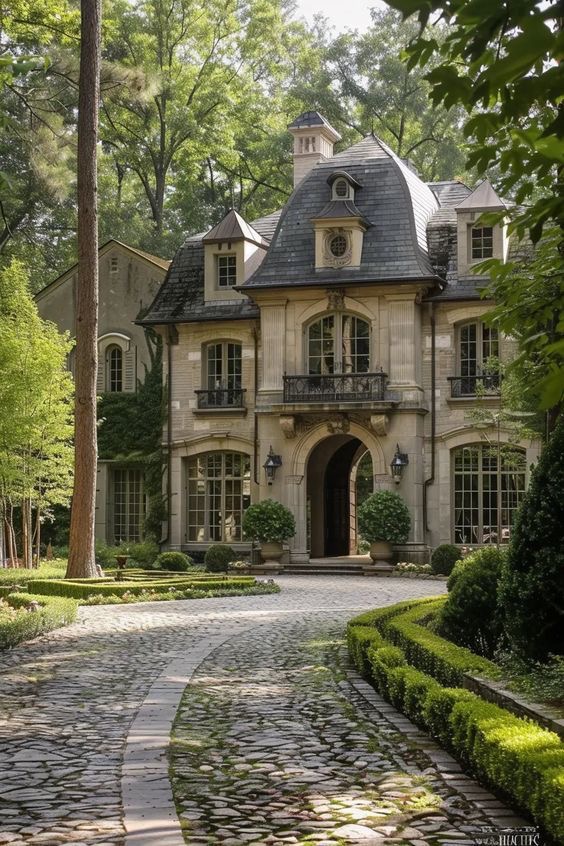 Elegant two-story mansion with cobblestone driveway, surrounded by lush greenery, featuring classic architectural details.