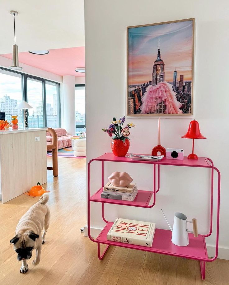 Modern living room with vibrant decor, pink shelf with books and decor, artwork of NYC skyline, and a pug walking on wood floor.