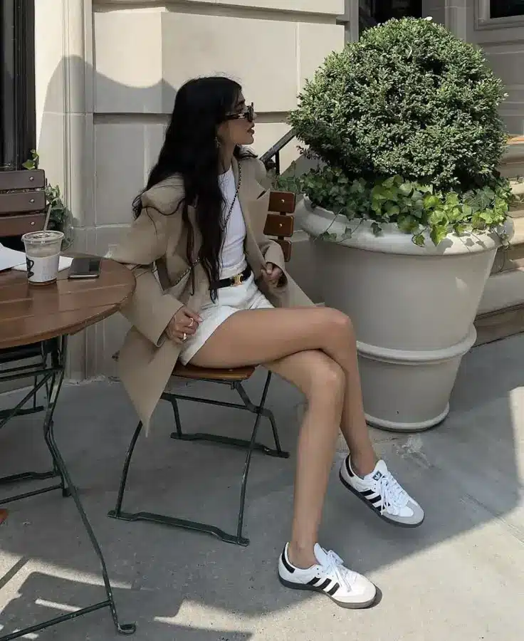 Woman sitting at a cafe in beige blazer, white shorts, and sneakers, with coffee and a plant nearby. Casual street style.