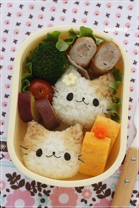 Cute cat-shaped rice balls in a bento box with broccoli, rolled meat, sweet potato, tomato, and tamagoyaki on a patterned cloth.