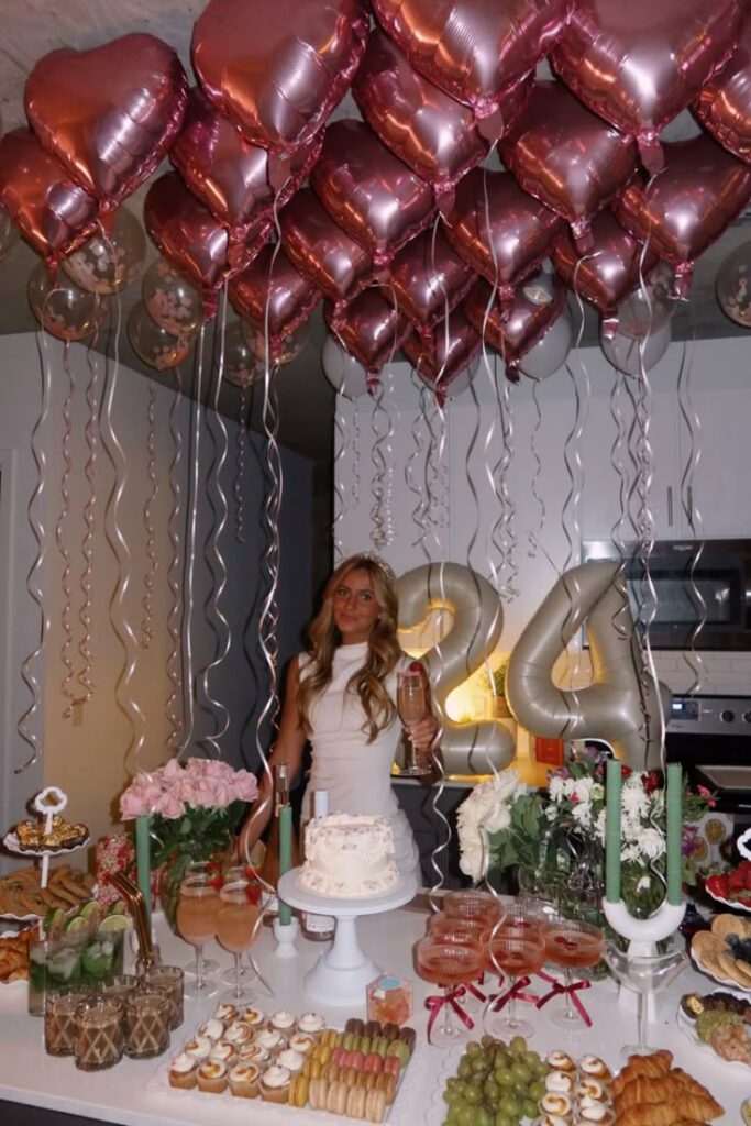 Birthday celebration with heart balloons, a woman in white, and a dessert table featuring a cake, flowers, and 24 balloons.