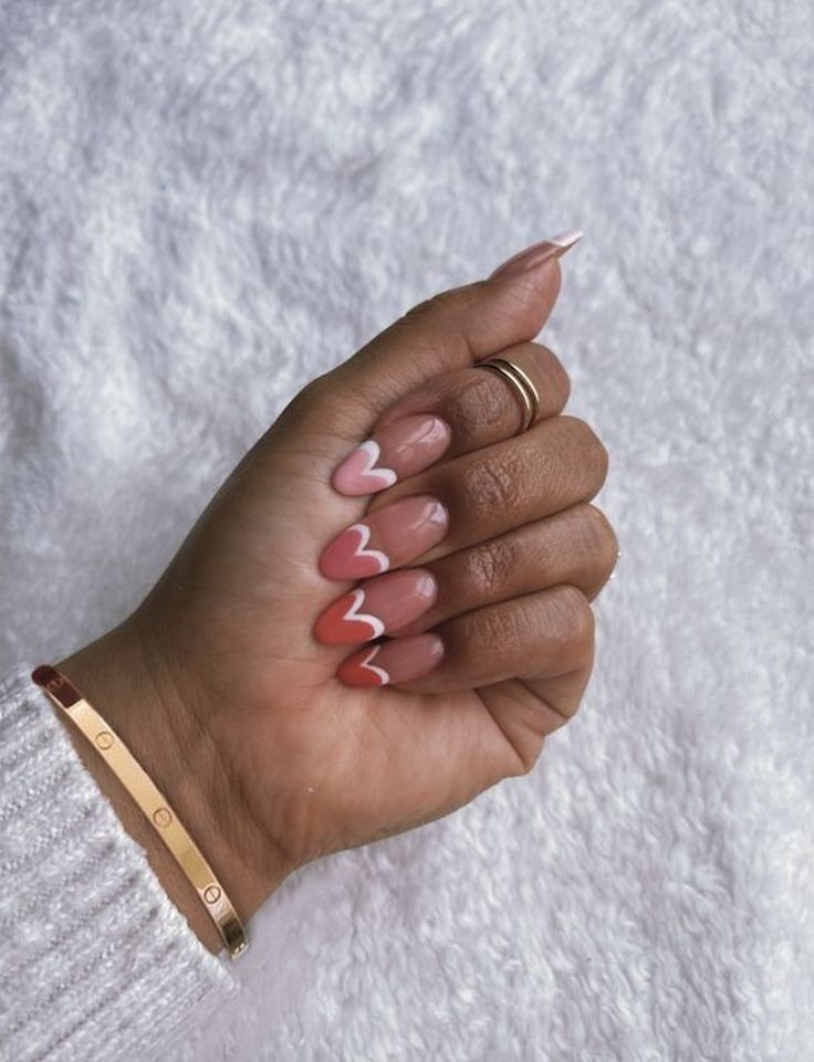 Hand with pink nail art and white heart designs, wearing gold rings and bracelet on a soft, white textured background.