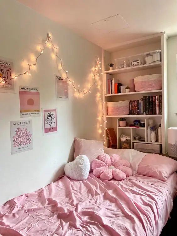 Cozy bedroom with pink decor, fairy lights, and pastel bedding. Bookshelf adorned with art prints completes the aesthetic look.