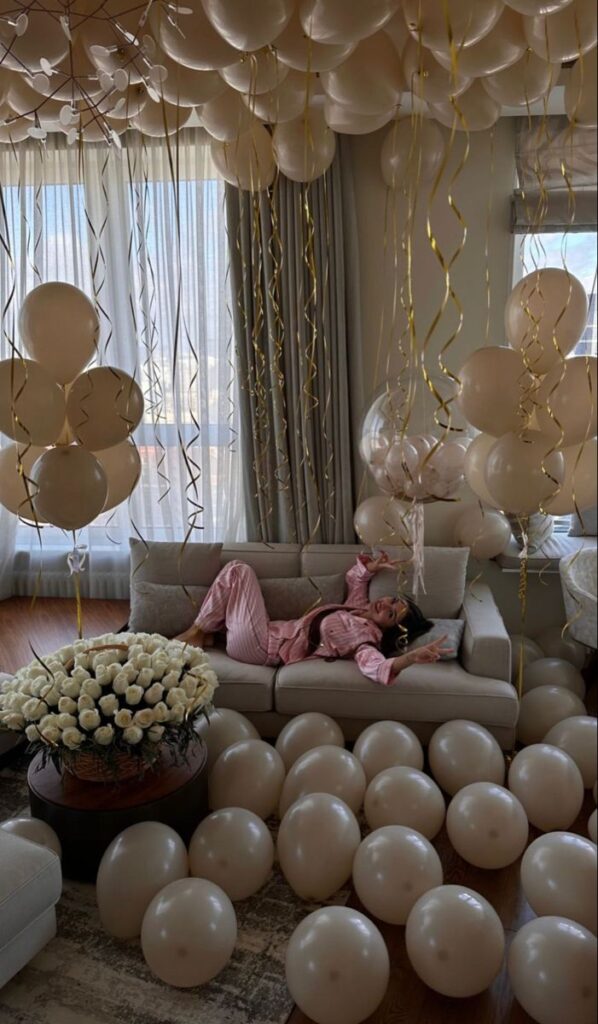 Woman in pink pajamas lounging on a sofa amidst cream balloons and white roses in a festive, light-filled room.