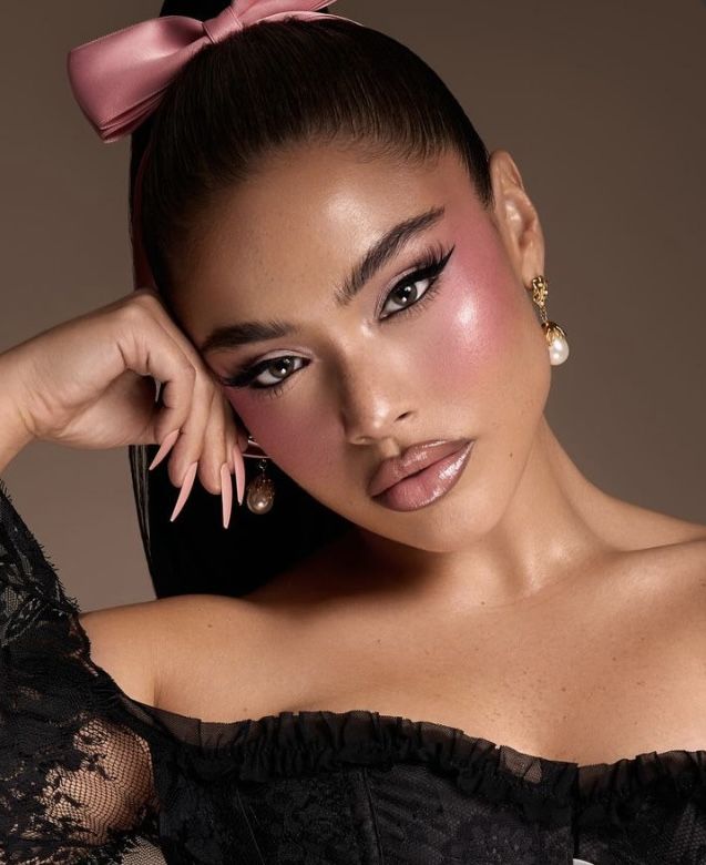 Woman with bold makeup, pink blush, pearl earrings, and pink bow headband, wearing lace outfit, gazing confidently.