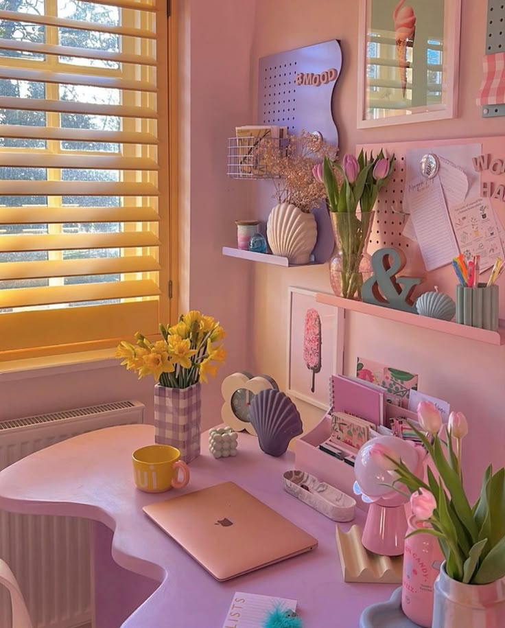 Pastel-themed home office with pink decor, tulips, and yellow vase, featuring a laptop on a curved desk by a sunny window.