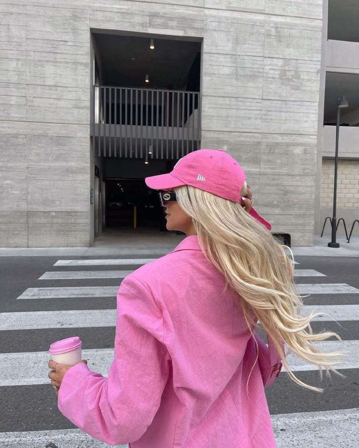 Woman in pink jacket and cap, holding coffee, walks across a city street with long blonde hair flowing behind her. Urban fashion style.