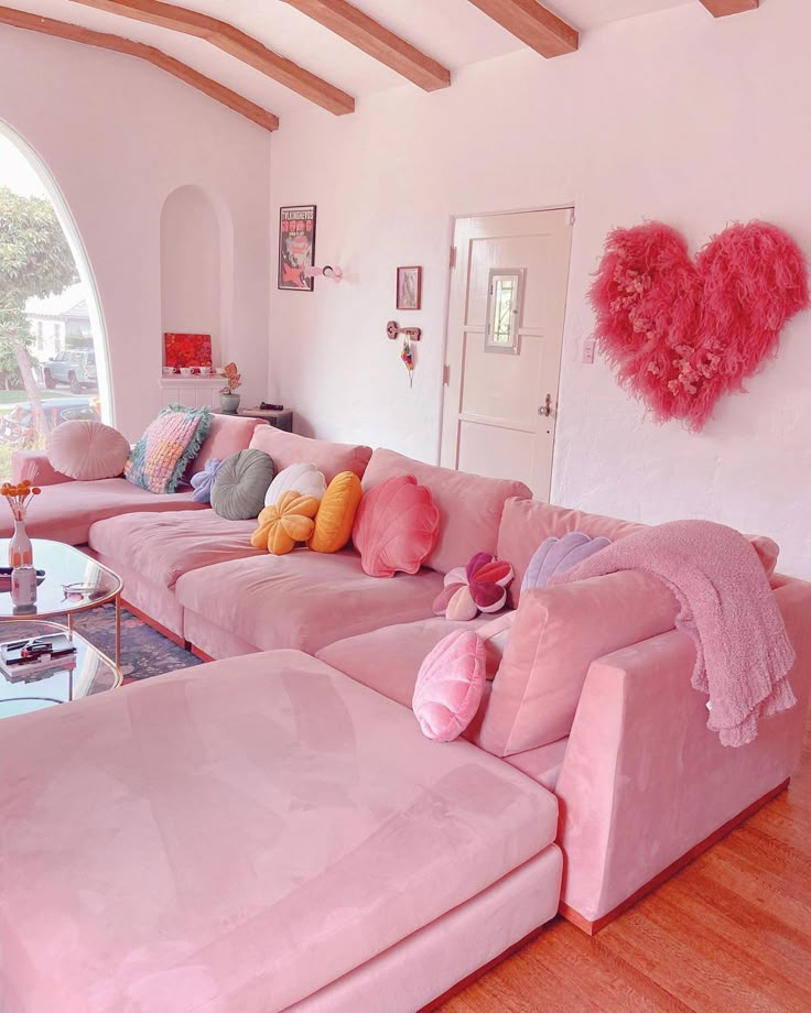 Cozy living room with pink decor: plush pink sofa, heart-shaped wall art, colorful cushions, and warm wooden accents.