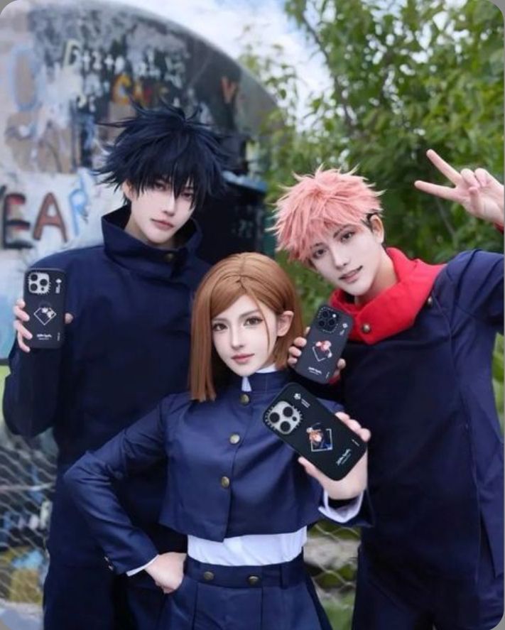 Cosplay trio in detailed anime costumes, holding phones with themed cases, outdoors with graffiti background and trees.