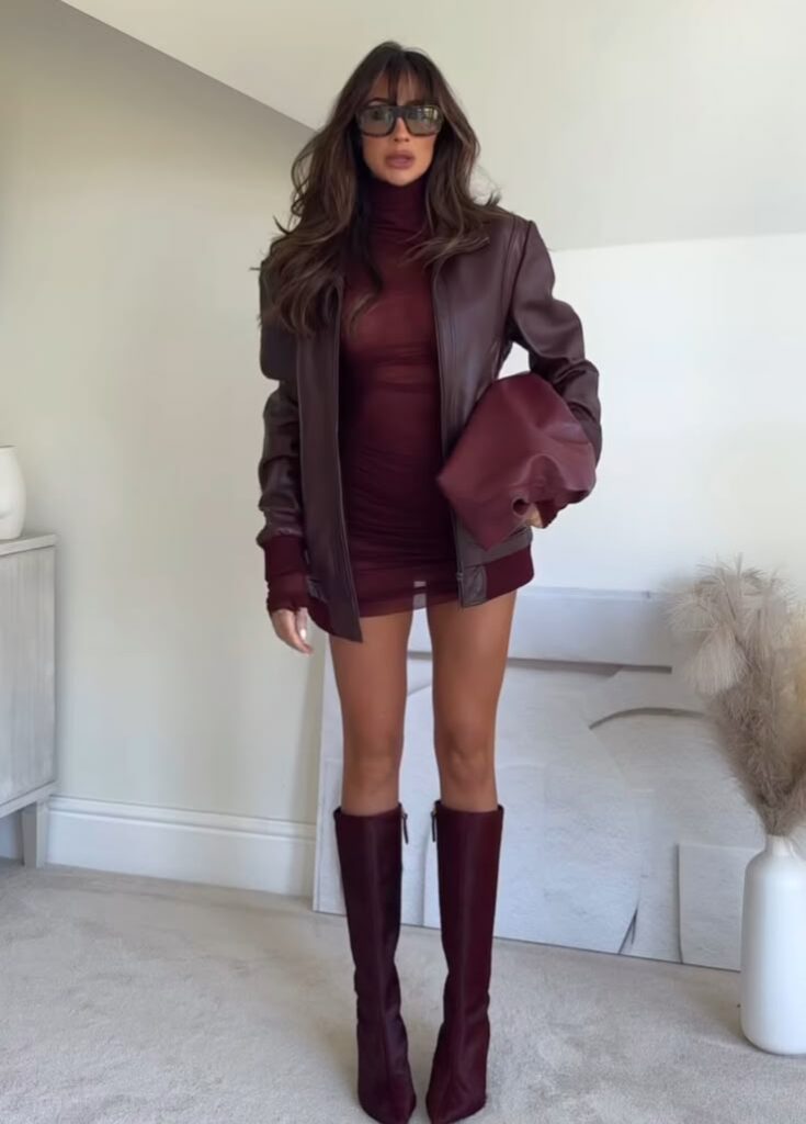 Fashionable woman in maroon mini dress with matching jacket, clutch, and knee-high boots, posing indoors. Trendy fall outfit.