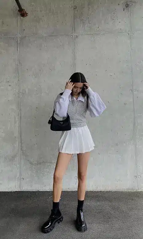 Fashionable woman in a pleated skirt, sweater, and boots, posing against a concrete wall, holding a black handbag stylishly.