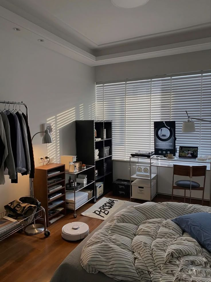 Cozy bedroom with striped bedding, shelves, a robot vacuum, and a stylish workspace bathed in natural light from blinds.