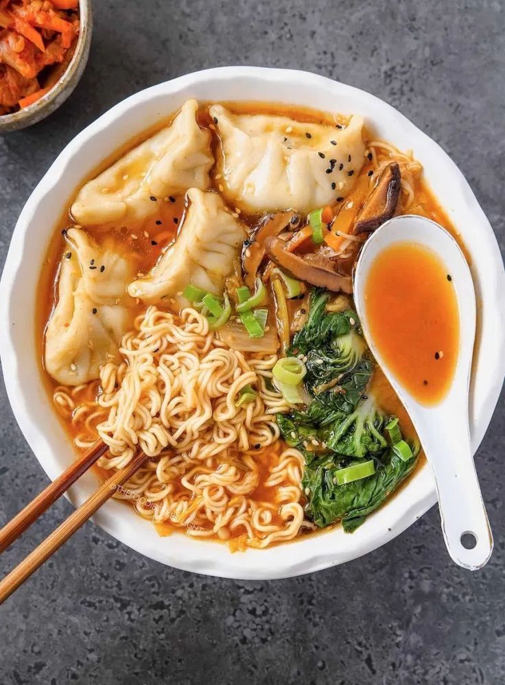 Bowl of flavorful ramen with dumplings, mushrooms, bok choy, and scallions in spicy broth, served with a spoon and chopsticks.