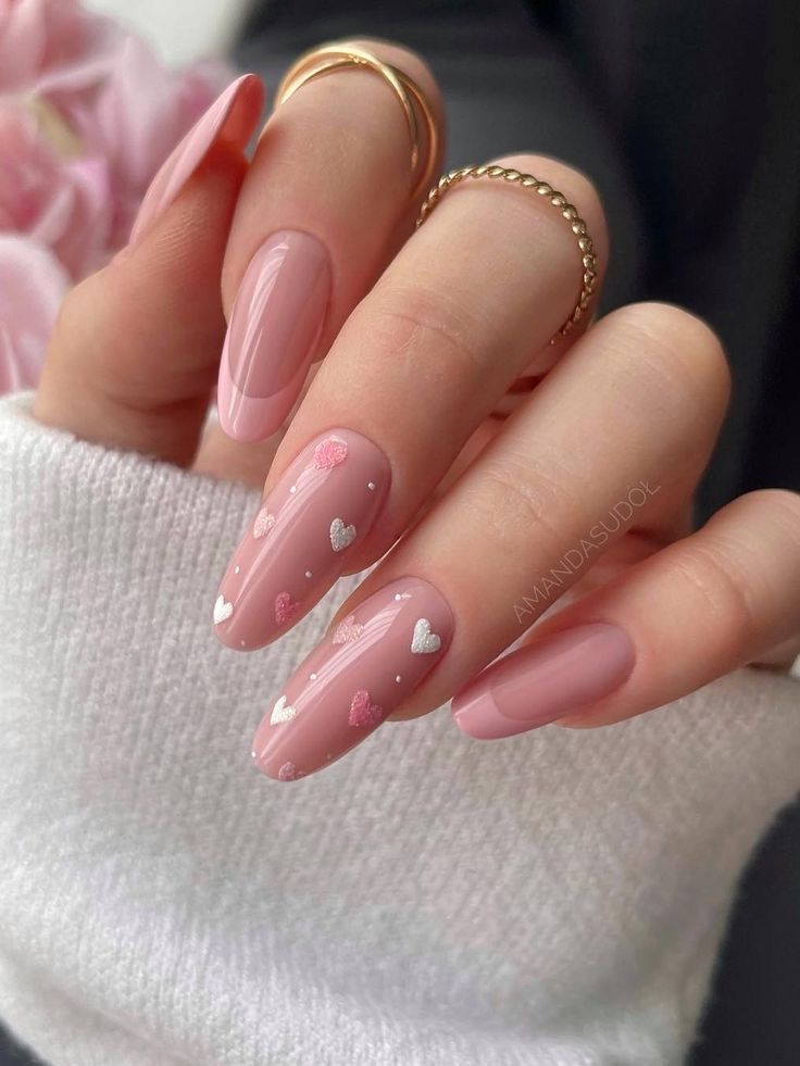 Long almond-shaped nails with pink polish and heart designs, adorned with gold rings on the hand. Fashionable nail art.