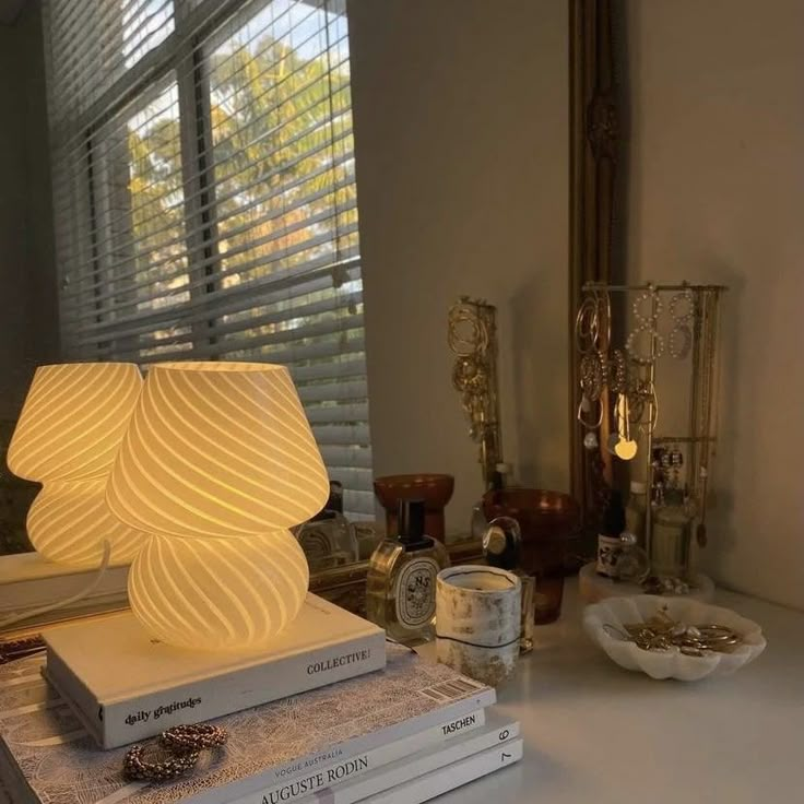 Cozy bedroom decor with glowing lamp on books, jewelry, and perfumes by a window with blinds. Calm and stylish ambiance.