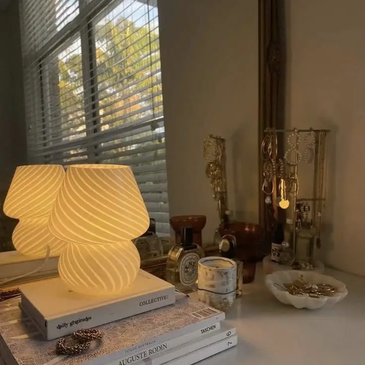 Cozy bedroom decor with glowing lamp on books, jewelry, and perfumes by a window with blinds. Calm and stylish ambiance.