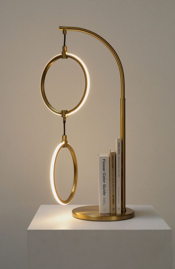 Modern brass lamp with two illuminated rings on a pedestal, next to books. Contemporary design and elegant home decor.