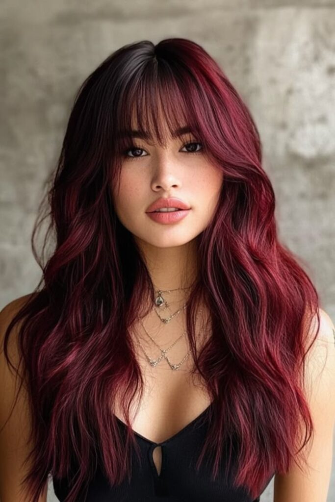 Woman with long, wavy dark red hair and bangs, wearing a black top and layered necklaces against a neutral background.