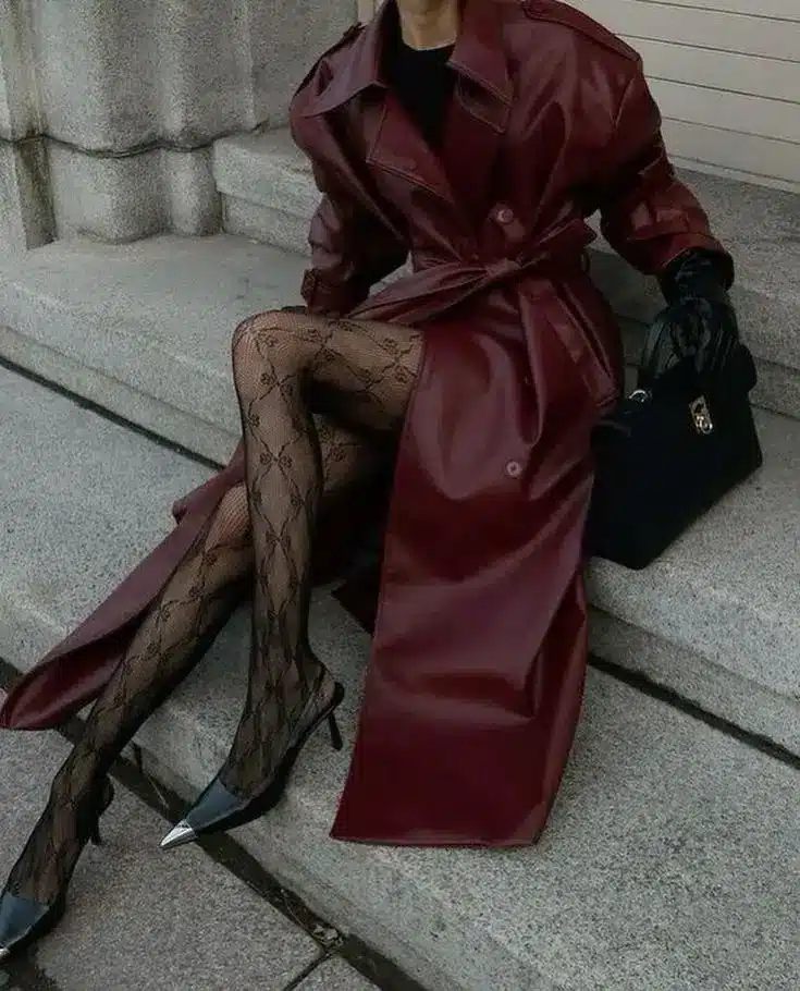 Woman in burgundy leather coat with patterned tights and pointed heels, holding a handbag on stone steps. Fashionable fall outfit.