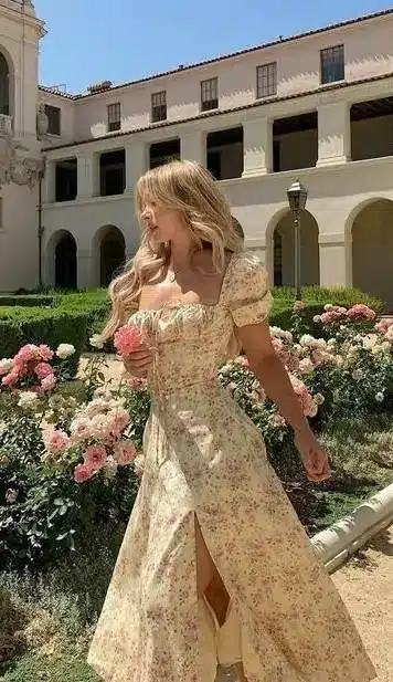 Woman in a floral dress admires roses in a sunlit garden, with elegant architecture in the background.