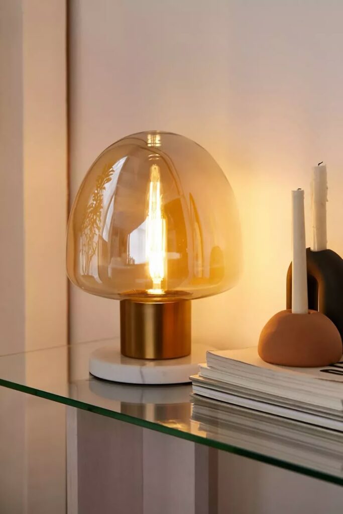 Modern amber table lamp on glass shelf, illuminating beside stacked books and decorative items. Cozy and stylish home decor.