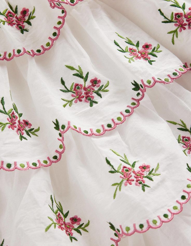 Intricate floral embroidery on white fabric with pink scalloped edges, featuring green leaves and pink flowers. Elegant textile design.