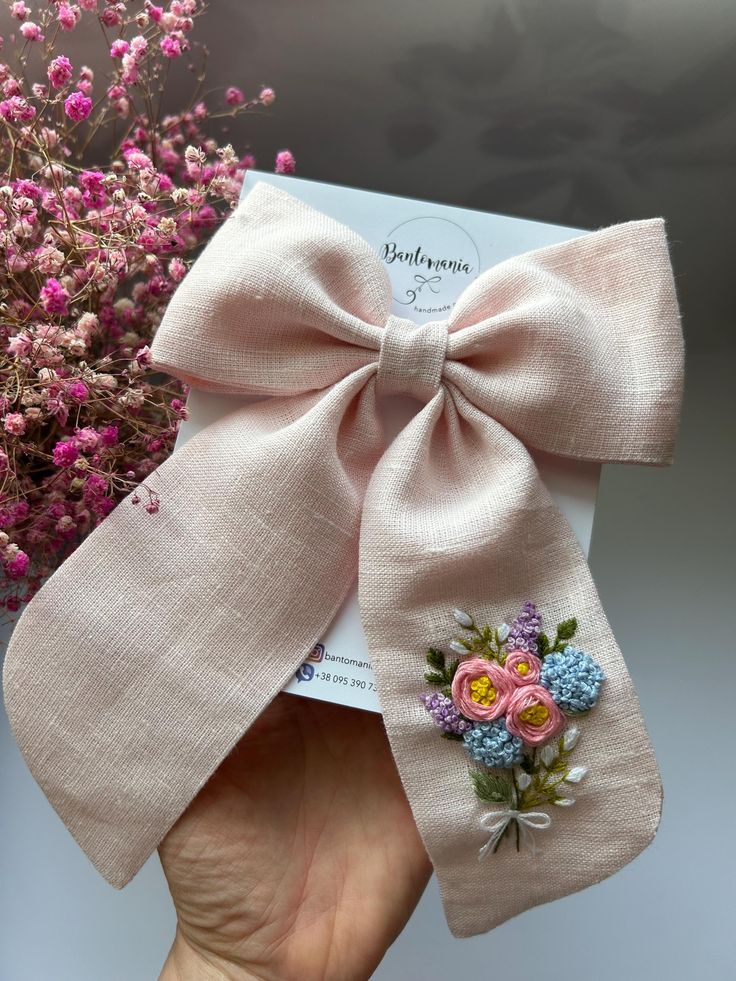 Handcrafted pink linen bow with floral embroidery, held by hand, set against a background of pink flowers.