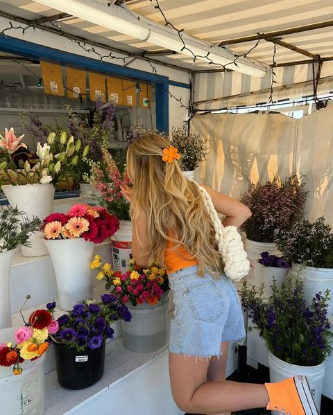 Woman with long hair, wearing an orange top and shorts, admires colorful flowers at a vibrant market stall.