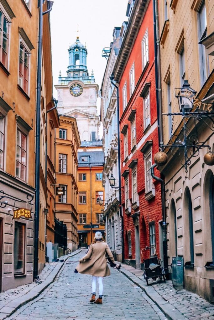 Woman exploring narrow cobblestone street with colorful buildings and clock tower in the background, vibrant urban travel scene.