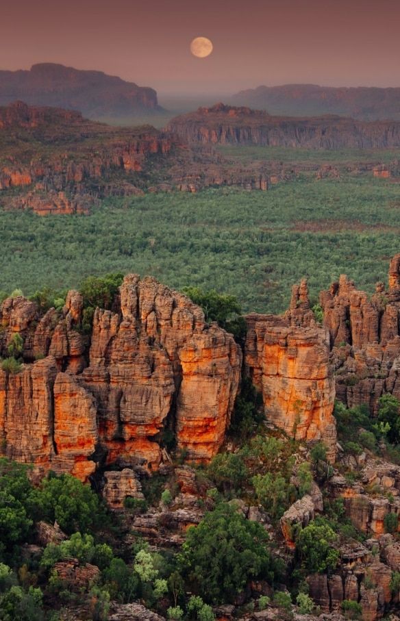 Moonrise over rugged red rock formations and lush greenery in a scenic mountainous landscape.