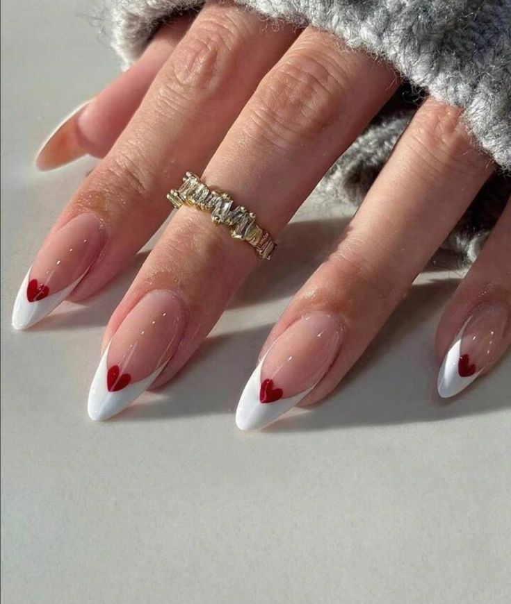 Elegant white French manicure with red heart accents, wearing a sparkling gold ring, on a soft gray fabric background.