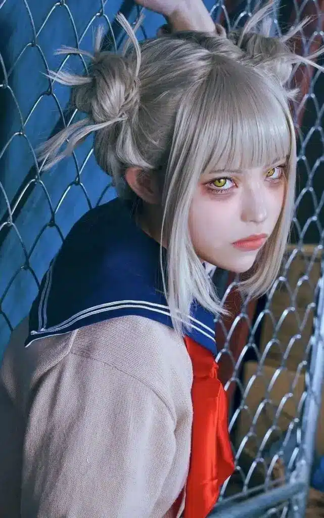 Anime girl with intense yellow eyes and silver hair in a bun, wearing a navy school uniform, stands by a chain-link fence.