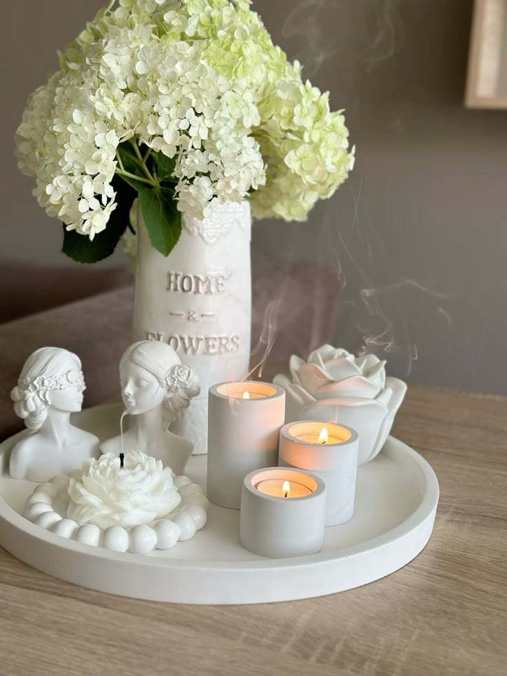 Elegant white floral arrangement with lit candles on a tray, featuring vintage-style busts and vase. Serene home decor setting.