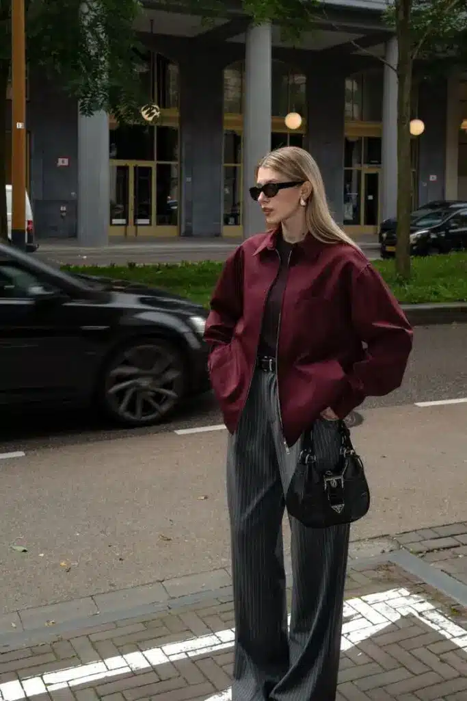 Fashionable woman in maroon jacket and striped pants standing on urban street, wearing sunglasses and holding a black handbag.
