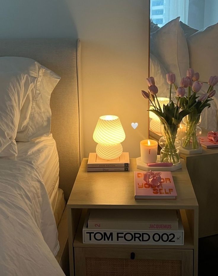 Cozy bedroom nightstand with a glowing lamp, pink tulips, candle, and fashion books, creating a warm, stylish ambiance.