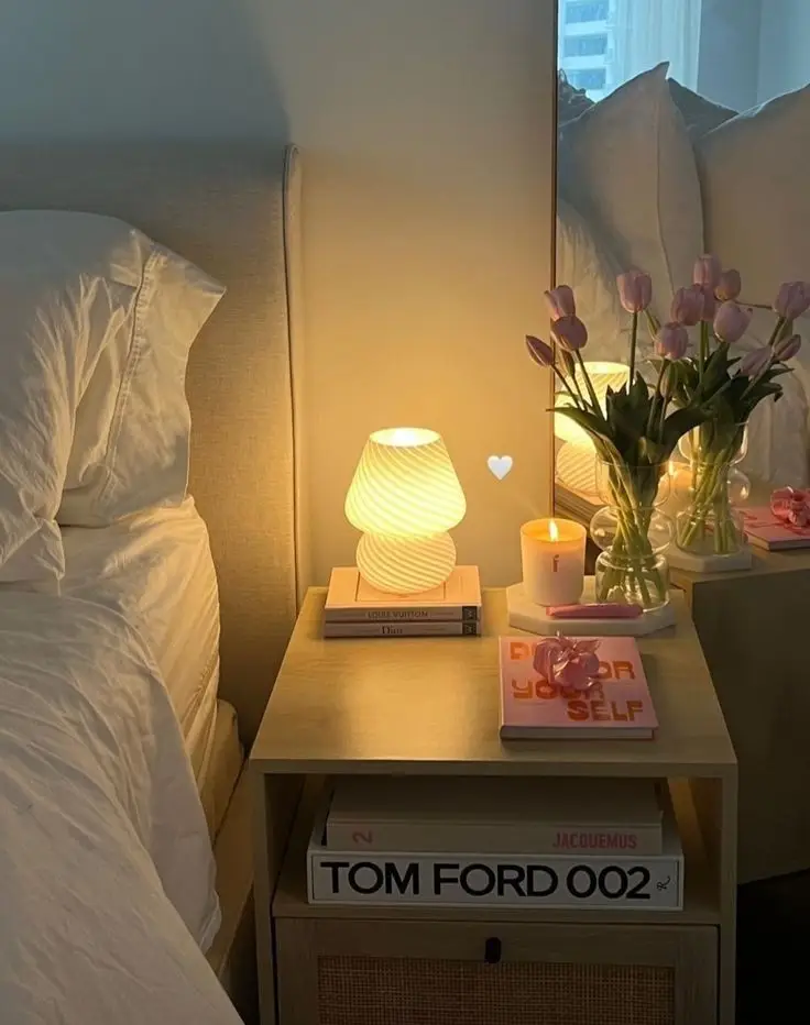 Cozy bedroom nightstand with a glowing lamp, pink tulips, candle, and fashion books, creating a warm, stylish ambiance.