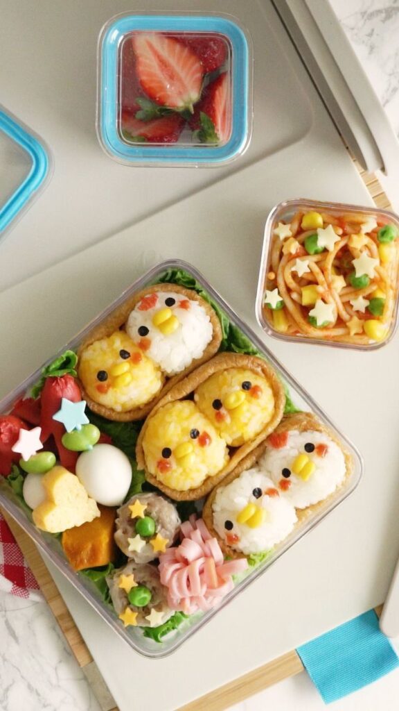 Cute bento lunch with rice shaped as chicks, pasta with vegetables, and fresh strawberries, perfect for a fun and creative meal.