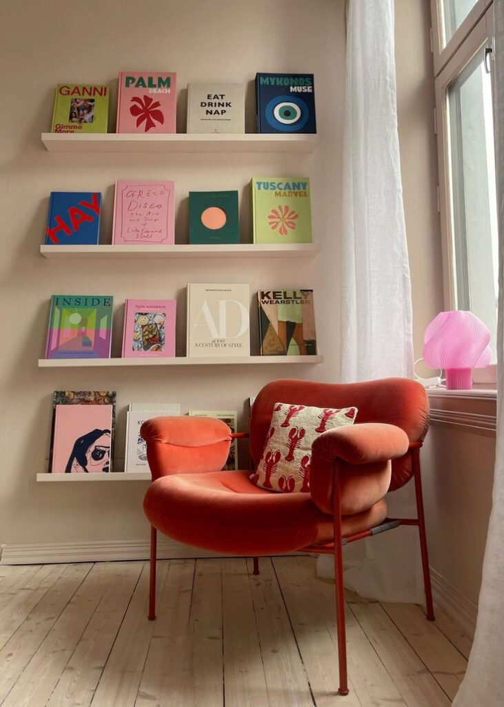 Cozy reading nook with modern orange chair, shelf of colorful books, and pink table lamp near a window with sheer curtain.