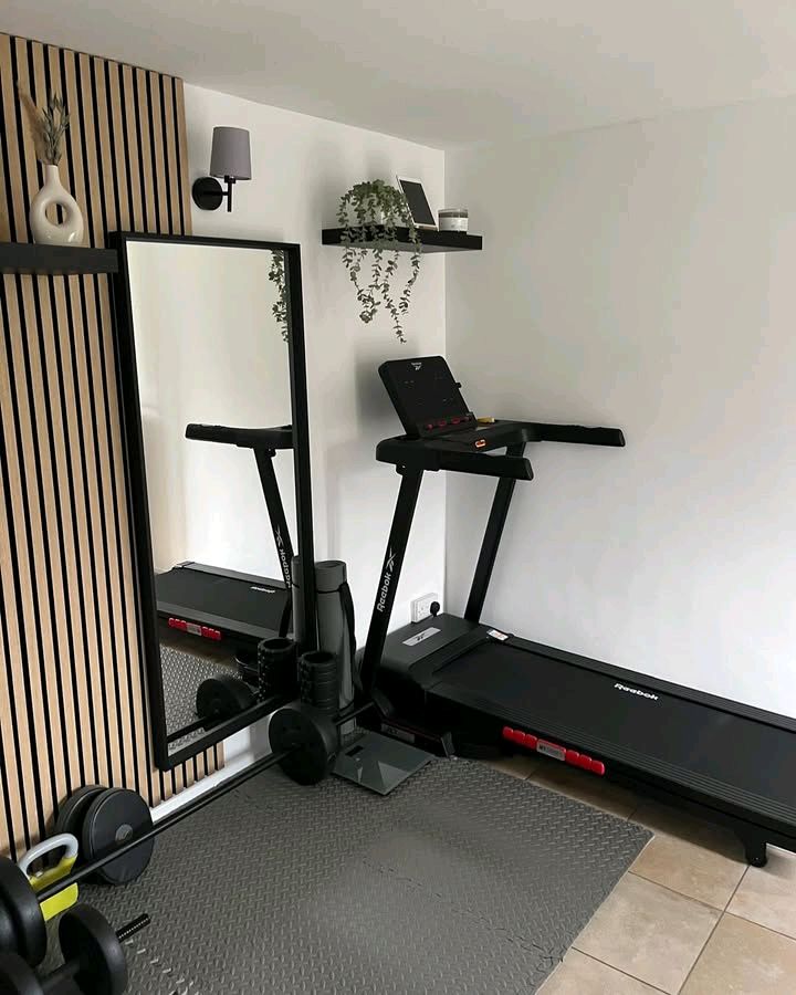 Home gym setup with treadmill, weights, and mirror on a mat. Modern decor with shelves, plant, and wall light. Ideal fitness space.