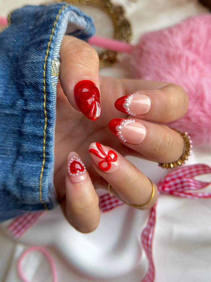 Stylish Valentine's Day nail art with red hearts, pearls, and bows on nude polish, held in a denim sleeve.