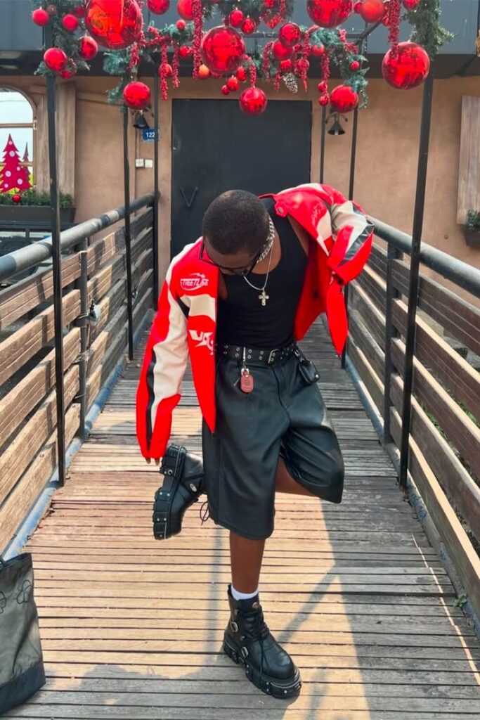 Fashionable person in red and black streetwear poses stylishly under festive decorations on a bridge.