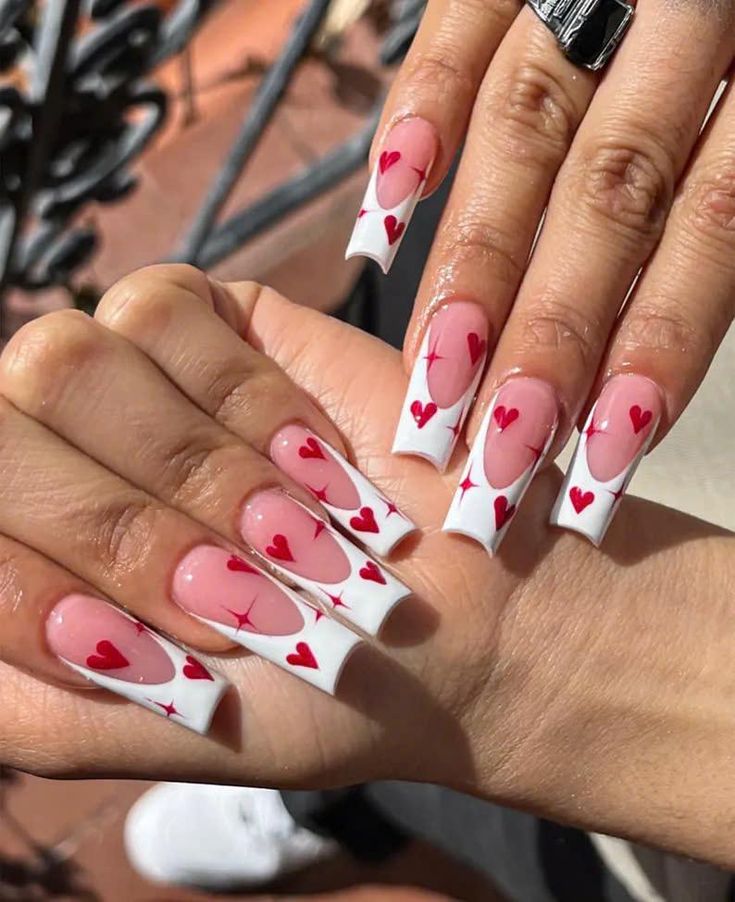 Long nails with pink and white French tips featuring red heart designs, showcasing a trendy and romantic manicure style.