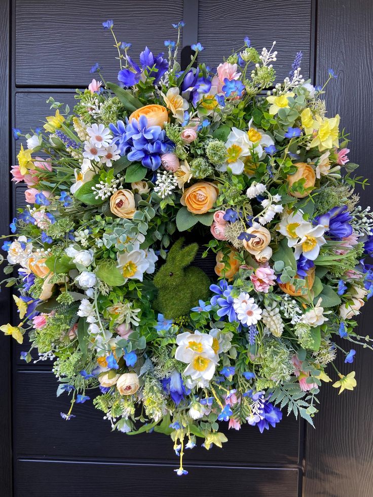 Vibrant floral wreath with a green rabbit centerpiece, featuring blue, yellow, and pink blossoms on a dark wooden door backdrop.