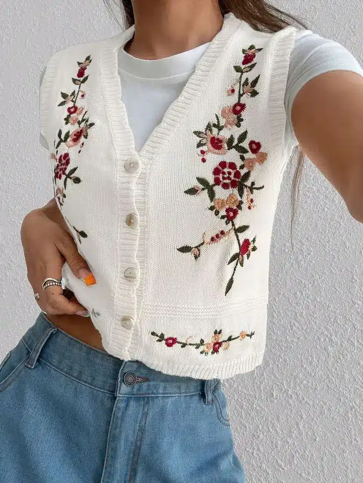 Woman in floral embroidered white vest and jeans, showcasing casual fashion style against a textured wall background.