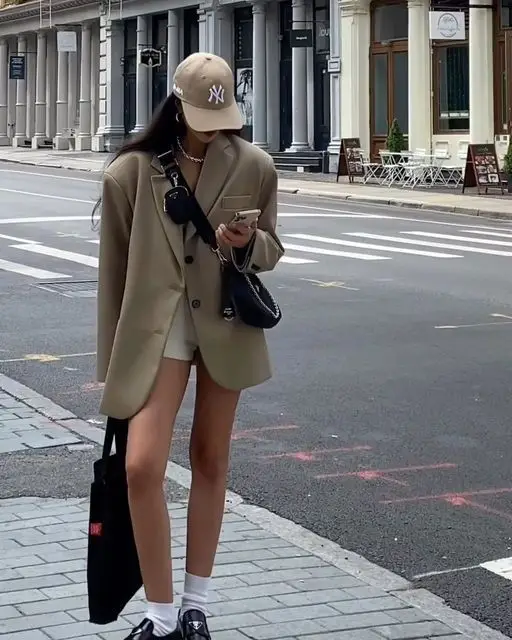 Fashionable woman in oversized blazer and cap, checks phone on urban street. Chic street style with crossbody bag and casual sneakers.