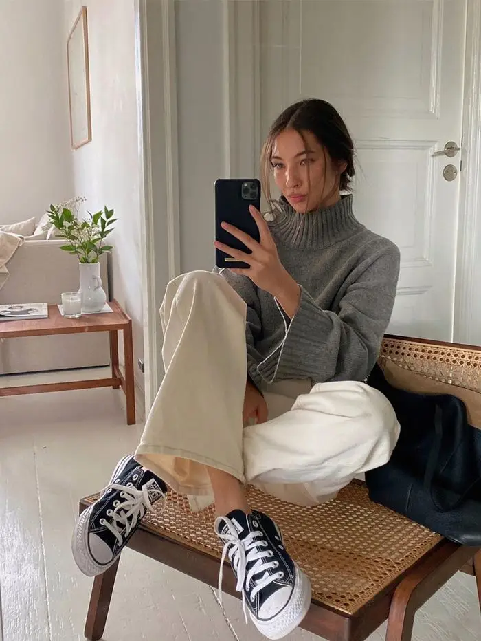 Woman in cozy grey sweater and beige pants taking a mirror selfie, casual style with black sneakers, chic interior background.