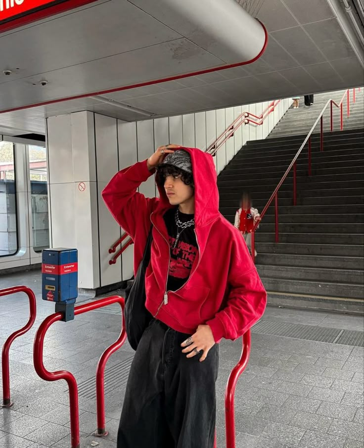 Person in red hoodie and black pants stands in a metro station, holding head, with stairs in the background. Urban fashion scene.