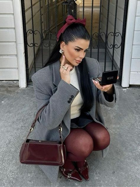 Fashionable woman in chic gray blazer applies makeup, accessorized with a burgundy bow, tights, and matching bag, outside a white building.